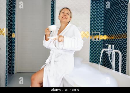 Eine junge Frau im weißen Bademantel trinkt Kaffee und nimmt ein Schaumbad. Die Blondine lächelt und genießt ein heißes duftendes Getränk. Lady ist ge Stockfoto