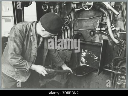 Dieses Foto zeigt einen Stoker der Nederlandse Spoorwegen in Aktion, der Kohle in den Feuerraum einer Dampfeisenbahn schaufelt. Sichtbare Elemente sind die Dampflokomotive, Kohle, Feuerwehrkasten, Schornstein, Räder, Pleuelstangen, Kessel, Heizer in Uniform, Werkzeuge, Kohlepfahl, Fabrikbahnhof, Außenbereich, mechanische Bauteile, Wartungsausrüstung, Bahngleise, und der niederländische Eisenbahnbetrieb Anfang des 20. Jahrhunderts, der die Praktiken des Dampflokomotivbetriebs veranschaulicht. Stockfoto