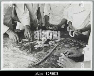 Foto von Bäckereiarbeiten, rollende Teigstränge für geflochtene Brote und Brezeln, Niederlande, 1940. Das Bild zeigt Bäcker, die Teig Formen, Knettische, zubereitete Teigstränge, traditionelle Brotbacktechniken, geflochtene Brote, Brezelbildung, Arbeitsumgebung, Backutensilien, kooperativer Arbeitsablauf in Bäckerei, Ofenvorbereitung, Mehlhandhabung, Teigkonsistenz, handwerkliches Können, niederländische Backmethoden, Bäckereipraxis des frühen 20. Jahrhunderts, Innenbäckerei-Layout, Teamarbeit, Backaktivität, Teigformung, kommerzielle Brotherstellung, Handel, Handwerk, berufliche Tätigkeit, professionelle Bäcker, Stockfoto
