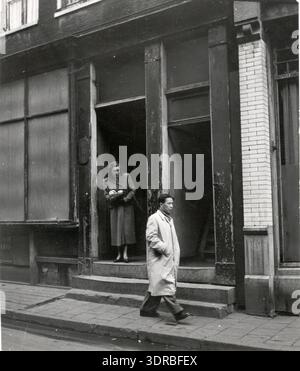 Im Jahr 1951 wird eine Frau fotografiert, die in einer Tür in Amsterdams Chinatown steht, während ein Chinese vorbeigeht. Das Bild zeichnet städtische Straßenlandschaft, Türarchitektur, Kleidung, Haltung, soziale Interaktion, Nachbarschaft, Gemeindeleben und chinesisch-holländische Kultur Mitte des 20. Jahrhunderts. Es dokumentiert das tägliche Leben, die menschliche Aktivität, die Präsenz auf der Straße und die kulturelle Umgebung in Amsterdam und bewahrt historische Beweise für persönliche und soziale Interaktionen. Stockfoto