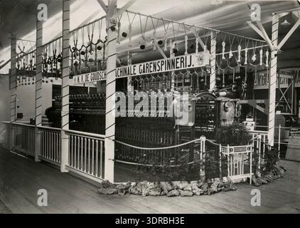 Dieses Foto von Kees Hofker zeigt die Wollweberei und die Herstellung von Decken am Garnspinnstand Goudse während einer Ausstellung in Gouda, Niederlande, 1913. Das Bild zeigt Webmaschinen, Spinnmaschinen, Decken, Garne, Arbeiter, die Maschinen bedienen, die Ausstellung zeigt Techniken der niederländischen Textilindustrie des frühen 20. Jahrhunderts, die maschinelle Produktion und die industrielle Arbeitspraxis. Stockfoto