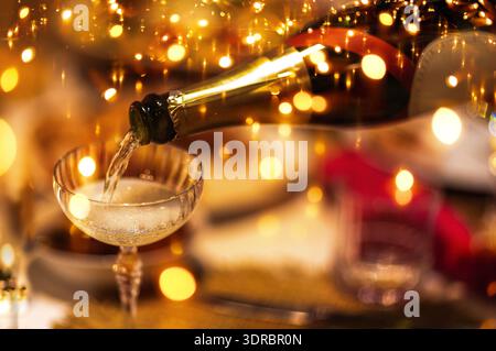 Champagner wird aus der Flasche in ein stilvolles Glas gegossen. Glitzernde goldene Festlichter vor Hintergrund. Blasen steigen in Glitzer auf Stockfoto