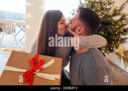 Ein glückliches verheiratetes Paar küsst sich unter einem geschmückten Weihnachtsbaum zu Hause. Ein attraktiver Mann schenkte seiner Schönheit ein Neujahrsgeschenk Stockfoto