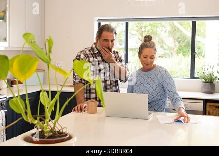 Ein älteres Ehepaar, das in der Küche einen Laptop benutzt, Dokumente liest und zu Hause Kaffee trinkt Stockfoto