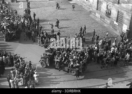 Am 8. Mai 1945 ist der Staudamm in Amsterdam in der Morgensonne voll von jubelnden Menschenmassen, als alliierte Militärfahrzeuge der Seaforth Highlanders of Canada in der Nähe des kaum sichtbaren Königspalastes vorbeifahren. Die Leute sind auf die Fahrzeuge geklettert und das Rote Kreuz ist anwesend. Fotografiert vom Pfarrhaus Nieuwe Kerk, zeigt das Bild städtische Feierlichkeiten, zivile Engagements und die Befreiung Amsterdams. Stockfoto