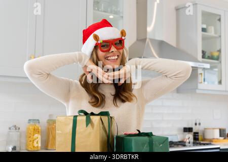 Lächelnde Frau im Weihnachtsmann-Hut posiert mit Weihnachtsgeschenken in der Küche, zu Hause Stockfoto