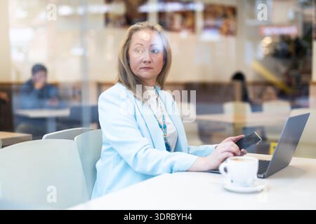 Geschäftsessen: Geschäftsfrau sitzt am Tisch im Café am Fenster mit einem Laptop und trinkt Kaffee. Ansicht durch Fenster Stockfoto