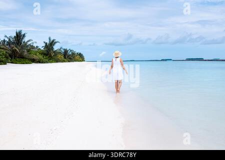 Eine junge Frau in weißem Kleid spaziert am Meer entlang. Die Aussicht von hinten. Eine Dame mit Strohhut genießt klares Wasser und Stockfoto