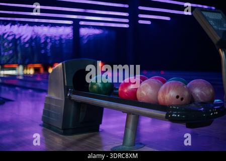 Bunte Bowlingkugeln liegen auf einem automatischen Rücklauf in einer dunklen, blau beleuchteten Gasse. Nahaufnahme von rosa und grünen Bowlingbällen in einem Unterhaltungszentrum u Stockfoto