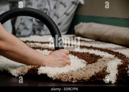 Ein Mann reinigt einen flauschigen, melierten Teppich mit einem schwarzen Staubsaugerschlauch. Die Hand Einer Person hält einen Staubsaugerschlauch während der professionellen Reinigung eines l Stockfoto