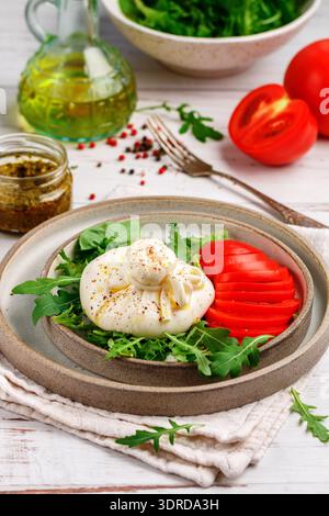 Cremiger italienischer Burrata-Käse serviert mit frischem Rucola, Tomaten, Olivenöl, Basilikum-Pesto und frisch gemahlenem Pfeffer auf dem Grau Stockfoto