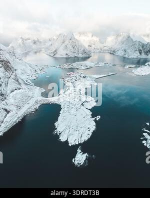 Aus der Vogelperspektive der schneebedeckten reine taucht im dunklen Wasser des Fjords auf, gekrönt von zerklüfteten Gipfeln, die den frostigen Himmel durchdringen, reine, Nordland, Norwegen. Stockfoto