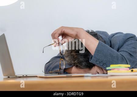 Ein junger Mann, der von Arbeitsstress überwältigt ist, legt seinen Kopf auf einen Schreibtisch und reflektiert Burnout, Ermüdung und modernen Druck am Arbeitsplatz. Stockfoto