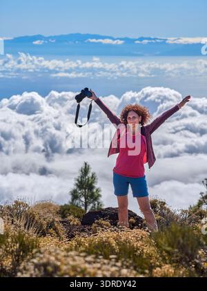 Eine fröhliche Frau mit lockigem rotem Haar hebt triumphierend die Arme, während sie eine Kamera hält, und steht über einem dramatischen Wolkenmeer an den Hängen des Teide Stockfoto