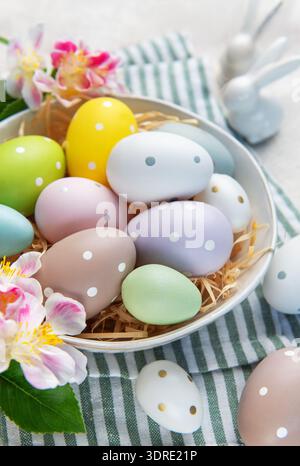 Pastellfarbene ostereier mit gepunkteten Mustern in einer Schüssel mit Strohhalm, Blumen und Hasenfiguren Stockfoto