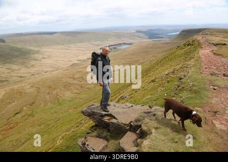 Ein lächelnder männlicher Wanderer mit großem Rucksack steht auf einem Felsvorsprung am Rande einer dramatischen Steilküste im Brecon Beacons National Park, Wales. Stockfoto