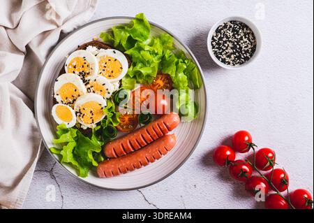 Gebratene Würstchen, Tomaten, Salat und ein Sandwich mit einem gekochten Ei auf einem Teller mit Blick von oben Stockfoto