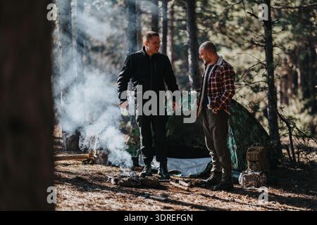 Zwei Männer campen in einem Kiefernwald mit einem rauchigen Lagerfeuer in der Nähe eines Tarnzelt Stockfoto