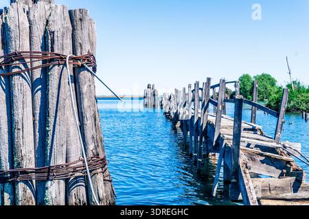 Ein hölzerner Pier mit einer Brücke über einem Gewässer. Die Brücke ist alt und verrostet, und der Pier ist zur Seite geneigt. Das Wasser ist ruhig und blau, und t Stockfoto