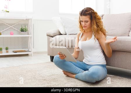 Frau sitzt auf dem Boden mit Tablet im Wohnzimmer und sieht verwirrt aus Stockfoto