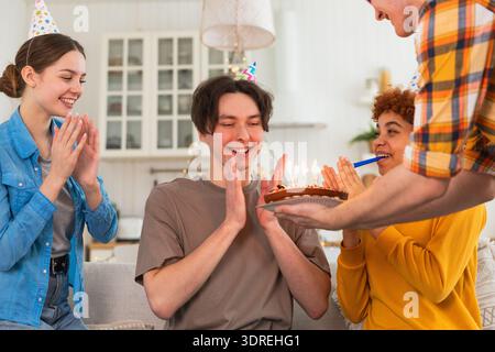 Wünschen Sie sich etwas. Mann mit Partymütze, der brennende Kerzen auf Geburtstagskuchen ausbläst. Happy Birthday Party. Gruppe von Freunden wünscht g Stockfoto
