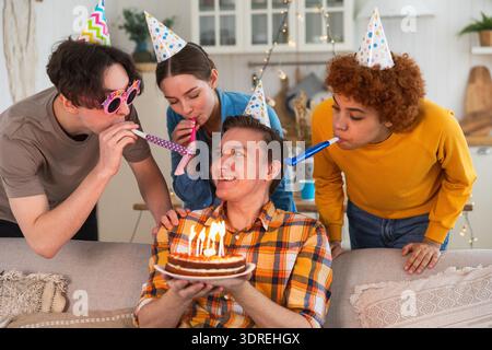 Wünschen Sie sich etwas. Mann mit Partymütze, der brennende Kerzen auf Geburtstagskuchen ausbläst. Happy Birthday Party. Gruppe von Freunden wünscht g Stockfoto