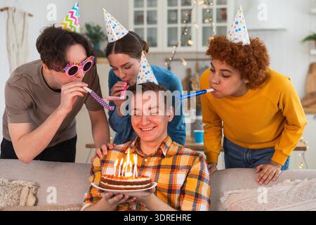 Wünschen Sie sich etwas. Mann mit Partymütze, der brennende Kerzen auf Geburtstagskuchen ausbläst. Happy Birthday Party. Gruppe von Freunden wünscht g Stockfoto