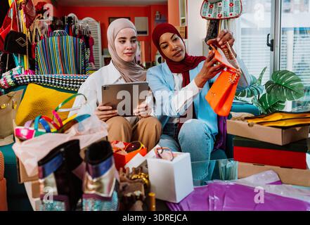 Zwei muslimische Unternehmerinnen sitzen zusammen, schauen sich eine stilvolle orange Handtasche an und diskutieren über neue Modeprodukte für ihre kleine Online-E-Kommunikation Stockfoto