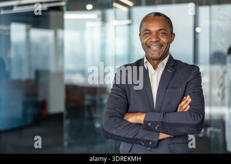 Ein selbstbewusster, reifer Geschäftsmann posiert mit einem freundlichen Lächeln und überkreuzten Armen in einem modernen Unternehmensumfeld und projiziert Erfolg und Führung Stockfoto