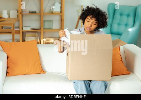 Afrikanisches Mädchen, das die Lieferung auspackt und in der Kiste sucht. Glückliche Frau, die den Karton öffnet. Eine Frau, die Pakete bekommt und die gelieferte Ware ansieht Stockfoto