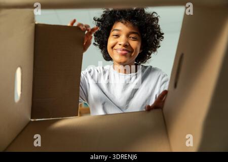 Innenseite des Kartons, Ansicht von unten. Afrikanisches Mädchen, das die Lieferung auspackt und in der Kiste sucht. Frau, die den Karton öffnet. Weibliche Paketbestellung Stockfoto