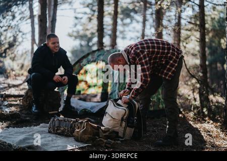 Zwei Männer campen in einem Wald, einer packt einen Rucksack in der Nähe eines Tarnzelt für Outdoor-Abenteuer Stockfoto