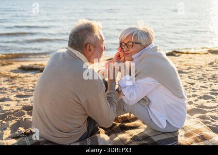 Älteres Ehepaar, das am Strand sitzt und Hände hält, um sich im Freien zu entspannen. Alte Ehefrau, die die Hände mit Zärtlichkeit berührt Stockfoto