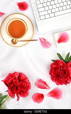 Moderner femininer Schreibtisch, Heimbüro. Computer, Smartphone, Pfingstrosenblüten und eine Tasse Tee auf einem sonnigen Tisch mit langen Schatten, Lifest Stockfoto