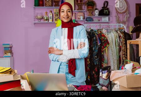 Selbstbewusste junge muslimische Unternehmerin, die mit überkreuzten Armen in ihrer kleinen Modeboutique steht und ihr Online-Geschäft und -Angebot präsentiert Stockfoto