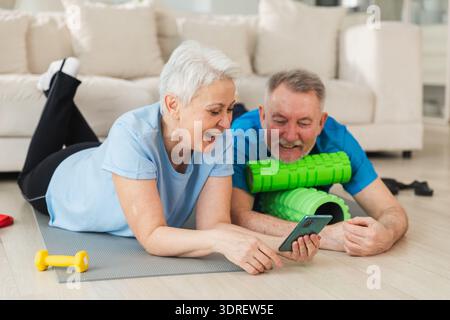 Yoga-Fitnesstraining online. Seniorenpaar üben Yoga-Fitness mit Online-Unterricht auf dem Smartphone. Die alte Ehefrau sieht zu Stockfoto