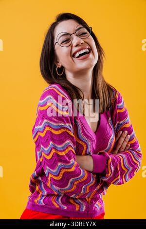 Junge Frau posiert selbstbewusst mit überkreuzten Armen, trägt einen bunten gestreiften Pullover und eine Brille auf einem leuchtend gelben Studiohintergrund und drückt fri aus Stockfoto