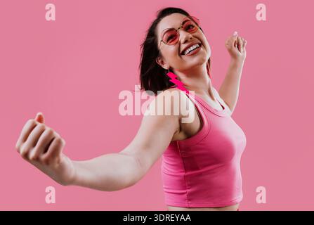 Glückliche junge Frau, die mit erhobenen Armen tanzt und Freude und Freiheit ausdrückt, während sie eine rosafarbene Sonnenbrille, ein Tanktop und rote Blitzohrringe trägt Stockfoto