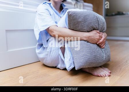 Nahaufnahme einer nicht erkennbaren Frau mittleren Alters, die das Kissen umarmt, während sie barfuß auf dem Schlafzimmerboden sitzt Stockfoto