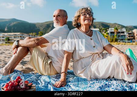 Älteres Ehepaar, das auf einer Stranddecke sitzt und sich den Rücken berührt, ein entspanntes Picknick mit Trauben und Feigen teilt, was für Ruhestand, Liebe und Leben steht Stockfoto