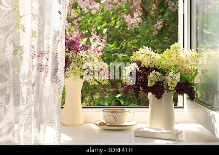 Stillleben mit blühenden Fliederblüten in einer Vase und einer Tasse Tee auf einem sonnigen Fenster, abstrakte Blumenarrangements, Inneneinrichtung Stockfoto