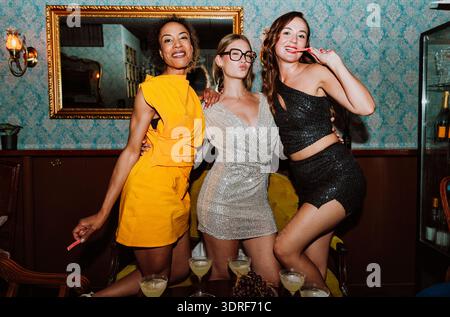 Drei verschiedene junge Frauen in Partykleidern posieren gemeinsam und feiern besondere Anlässe mit Cocktails und Snacks in einer stilvollen Bar oder Lounge Stockfoto
