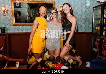 Drei verschiedene junge Frauen in Partykleidern posieren gemeinsam und feiern besondere Anlässe mit Cocktails und Snacks in einer stilvollen Bar oder Lounge Stockfoto