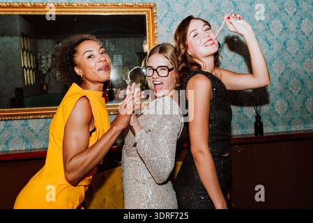 Drei verschiedene junge Frauen in Partykleidern posieren gemeinsam und feiern besondere Anlässe mit Cocktails und Snacks in einer stilvollen Bar oder Lounge Stockfoto