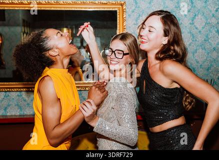 Drei verschiedene junge Frauen in Partykleidern posieren gemeinsam und feiern besondere Anlässe mit Cocktails und Snacks in einer stilvollen Bar oder Lounge Stockfoto