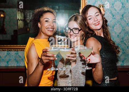 Drei verschiedene junge Frauen in Partykleidern posieren gemeinsam und feiern besondere Anlässe mit Cocktails und Snacks in einer stilvollen Bar oder Lounge Stockfoto
