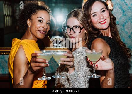 Drei verschiedene junge Frauen in Partykleidern posieren gemeinsam und feiern besondere Anlässe mit Cocktails und Snacks in einer stilvollen Bar oder Lounge Stockfoto