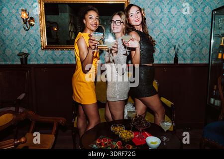 Drei verschiedene junge Frauen in Partykleidern posieren gemeinsam und feiern besondere Anlässe mit Cocktails und Snacks in einer stilvollen Bar oder Lounge Stockfoto