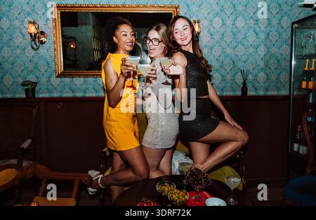 Drei verschiedene junge Frauen in Partykleidern posieren gemeinsam und feiern besondere Anlässe mit Cocktails und Snacks in einer stilvollen Bar oder Lounge Stockfoto