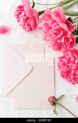 Pfingstrose Blumen und leere hellrosa Papierkarte mit offenem Umschlag auf weißem Leinentischtuch Hintergrund. Einladung Blumenkarte oder Brief in Vinta Stockfoto